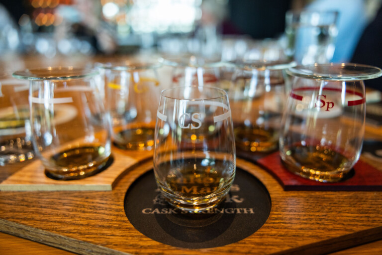 A wooden tasting tray featuring several whiskey tasting glasses, each engraved with letters and filled with amber liquid, set on a bar table.