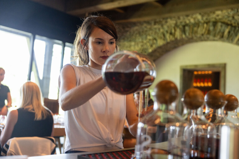 A woman in a sleeveless white top pours red liquid from a large glass vessel into smaller glass containers at a bar, with a rustic stone wall and several patrons in the background.