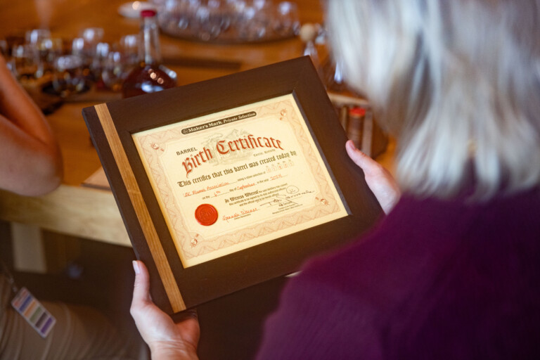 A person holding a framed 'Barrel Birth Certificate' from Maker's Mark, which certifies the creation of a barrel, featuring decorative elements and a red seal, with a soft-focus background of a wooden table and glasses.