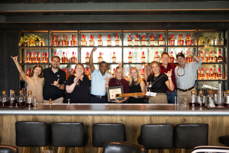 A group of ten individuals stands together in a bar setting, smiling and celebrating, with several holding glasses and a wooden plaque. Behind them, a wall is lined with bottles of bourbon. The atmosphere is festive and welcoming.