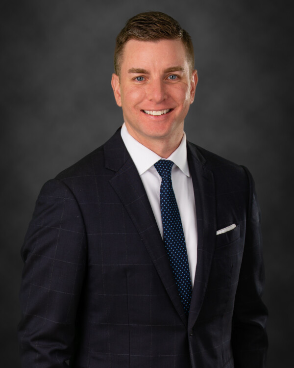A smiling man in a dark suit with a checked pattern and a blue tie featuring white polka dots, standing against a dark gray background.