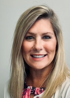 A smiling woman with long blonde hair, wearing a pink floral blouse and a cream-colored sweater, is posing for a close-up portrait against a light gray background.