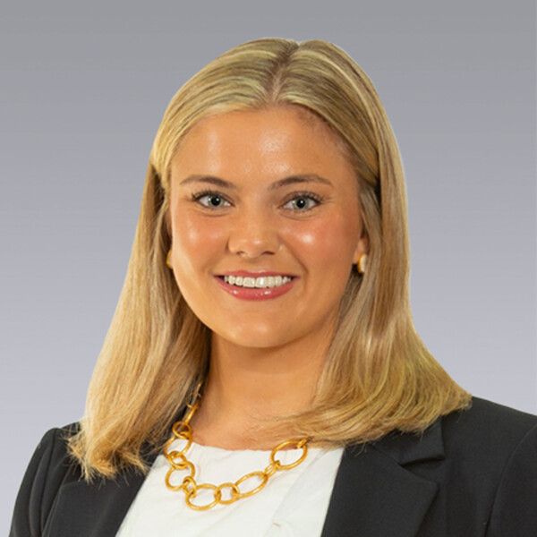 A young woman with shoulder-length blonde hair smiles at the camera, wearing a white blouse and a black blazer, accessorized with a gold chain necklace; the background is a soft gradient.
