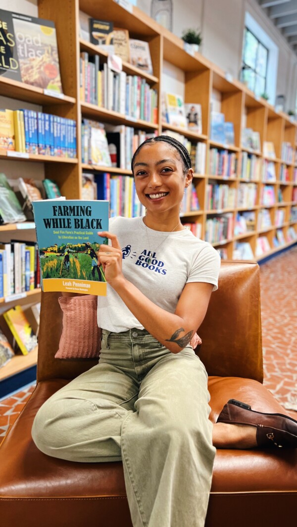 A person sitting on a brown leather chair in a bookstore, smiling while holding a book titled 'Farming While Black' by Leah Penniman. They are wearing a white t-shirt with 'ALL GOOD BOOKS' printed on it and light green pants, with braided hair and a tattoo visible on their arm. In the background, bookshelves filled with various books are visible.