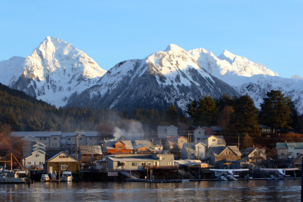 A picturesque waterfront scene featuring several wooden houses and boats along a river, with snow-capped mountains in the background under a clear blue sky.