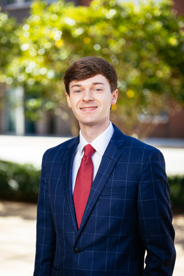A young man with short brown hair and a confident smile is standing outdoors, dressed in a navy blue blazer with a checkered pattern and a red tie. The background features blurred green foliage and urban architecture.