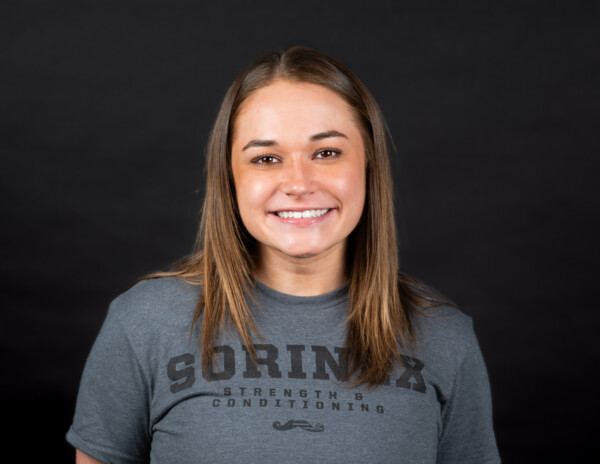 A smiling young woman with long brown hair wearing a gray t-shirt featuring the text 'SORINEX STRENGTH & CONDITIONING.' She is posed in front of a black background.