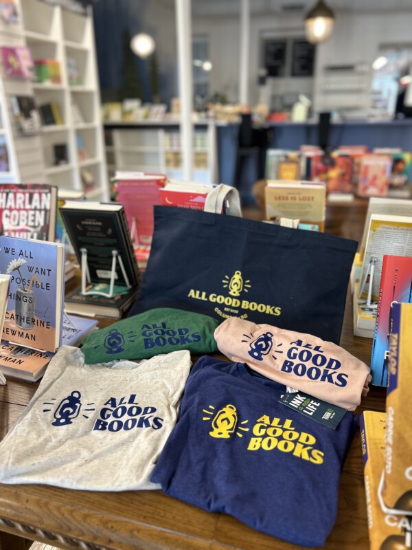 A selection of t-shirts and a tote bag featuring the text 'ALL GOOD BOOKS' displayed on a wooden table in a bookstore, surrounded by colorful book covers in the background.