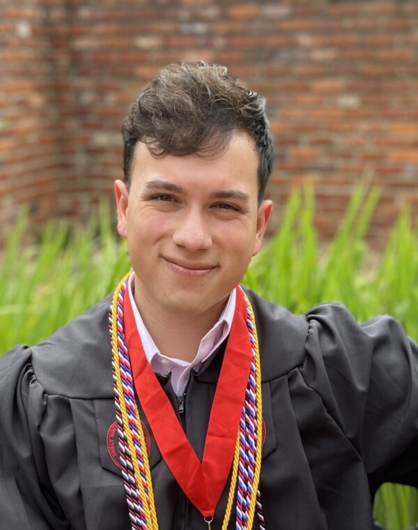 A young man in a black graduation gown and multiple honor cords sits on a decorative bench, smiling confidently with one leg crossed over the other, revealing colorful, patterned socks and brown dress shoes, surrounded by green grass and brick walls.