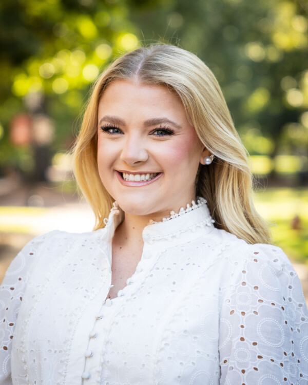 A smiling young woman with long blonde hair, wearing a white, intricately detailed blouse with pearl buttons, stands outdoors with blurred greenery in the background.