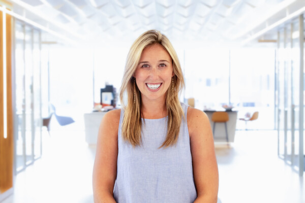 A smiling woman with long blonde hair wearing a sleeveless light blue top stands in a well-lit modern office space, with blurred furniture and natural light in the background.