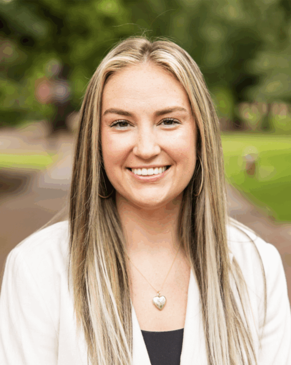 A young woman with long, straight blonde hair smiles at the camera, wearing a white blazer over a black top, against a blurred background of greenery and a pathway.