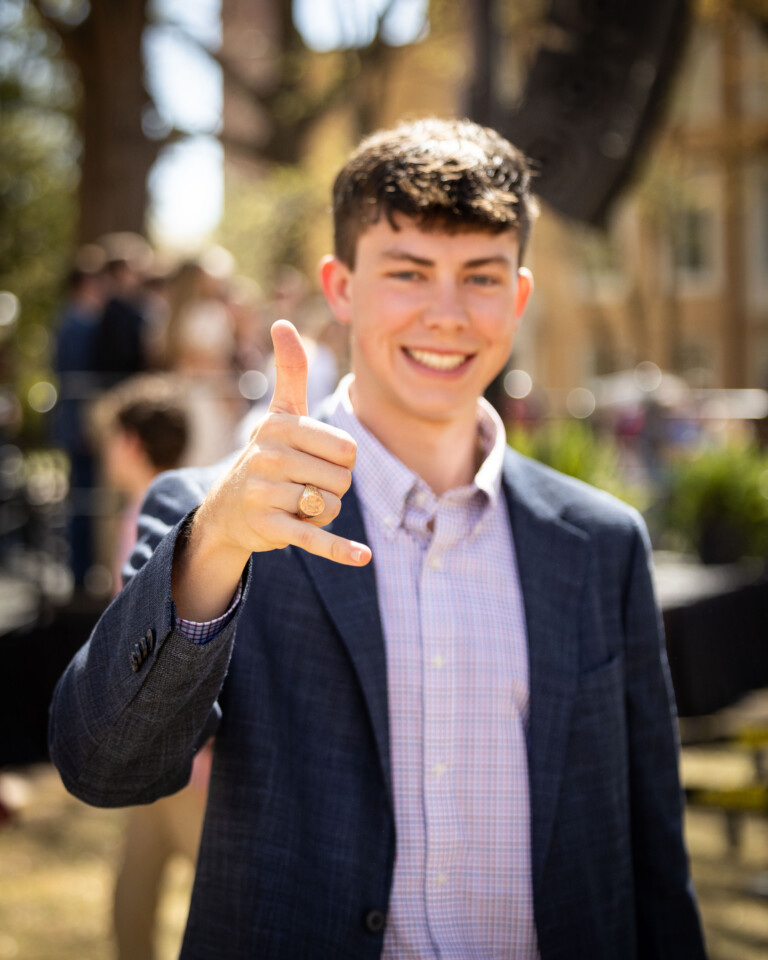 A young man in a navy blazer and a checkered shirt gives a thumbs-up gesture with one hand while smiling at the camera, with blurred people and greenery in the background.