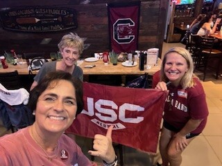 Three women posing for a selfie in a restaurant, with a USC flag in front of them and South Carolina memorabilia on the walls behind.