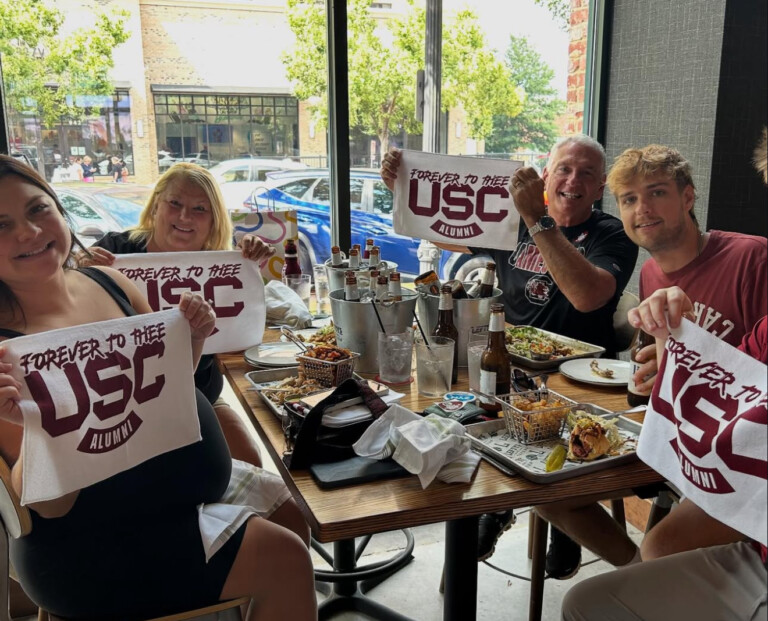 A group of four people sitting at a table inside a restaurant, smiling and holding white towels that read "FOREVER TO THEE USC ALUMNI" in maroon lettering. The table is filled with food, drinks, and various condiments, with windows showing a street view outside.