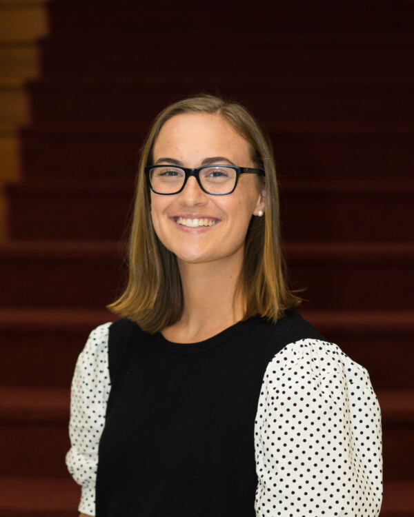 A smiling young woman with shoulder-length brown hair, wearing glasses and a black top with white polka dot sleeves, stands in front of a staircase with reddish carpet.