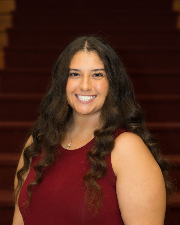 A woman with long, wavy brown hair smiles brightly, wearing a maroon sleeveless top, against a backdrop of soft red stairs.