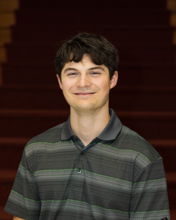 A young man with short, wavy brown hair smiles at the camera while wearing a grey and black striped polo shirt. He is positioned in front of a staircase with red carpet.