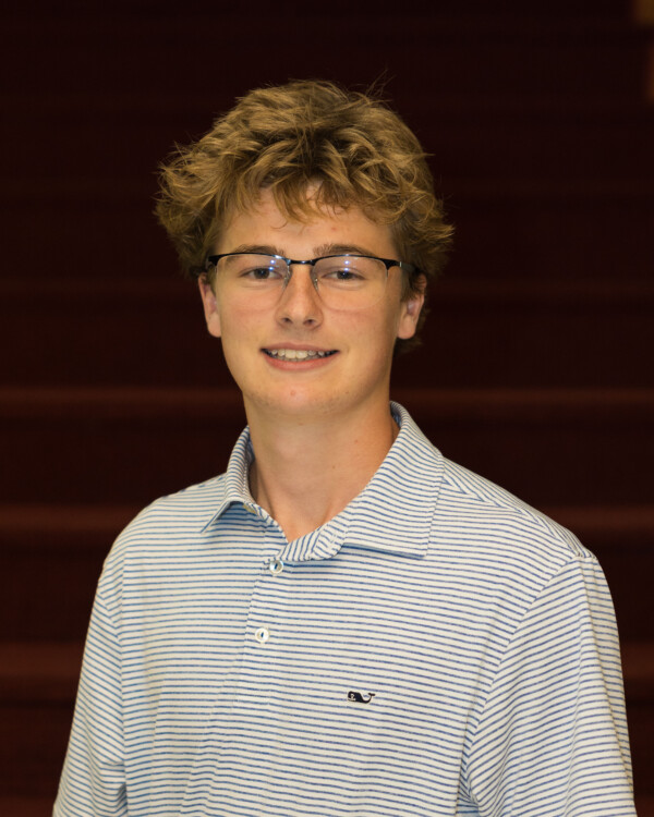 A young man with tousled blond hair and glasses smiles at the camera, wearing a blue and white striped polo shirt, with a softly lit background featuring steps.