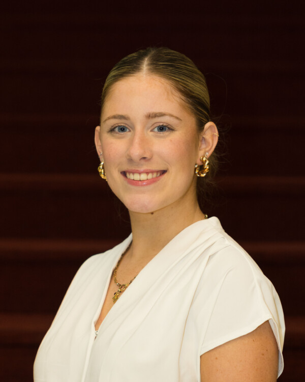 A woman with blonde hair styled back, wearing a white blouse and gold hoop earrings, smiles warmly in a portrait setting with a dark red background.