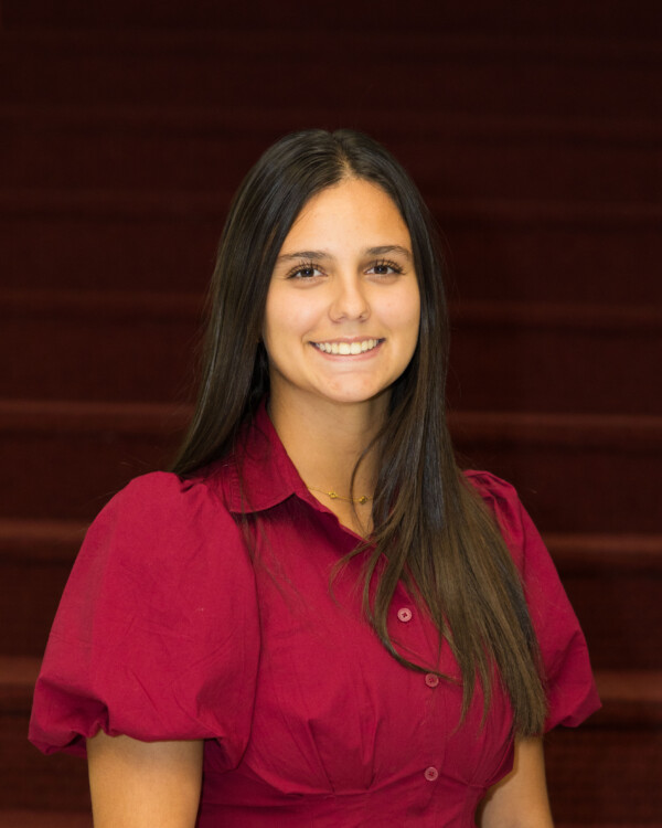 A young woman with long dark hair is smiling while wearing a short-sleeved, bright red blouse. She stands in front of a blurred background with staircase steps in maroon color.