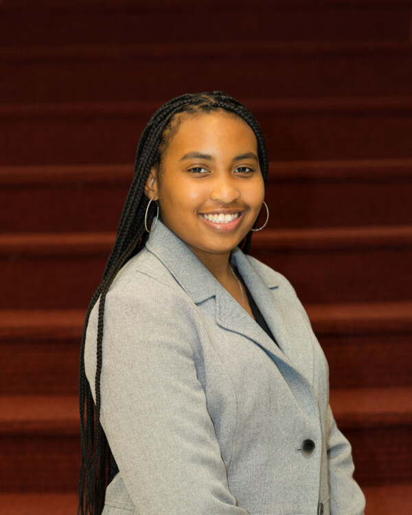 A young woman with braided hair wearing a light gray blazer smiles confidently against a backdrop of red-carpeted stairs.