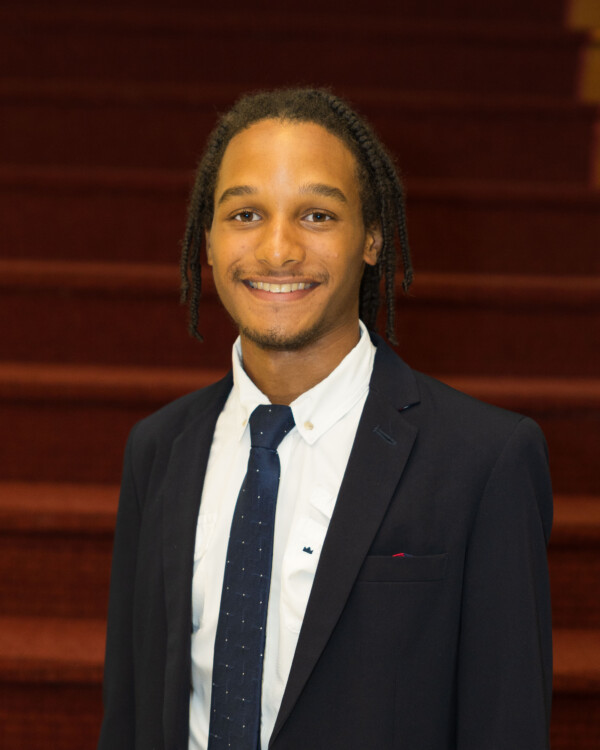 A young man with braided hair, smiling confidently, is dressed in a professional dark suit and tie, standing in front of a staircase with red carpeting.