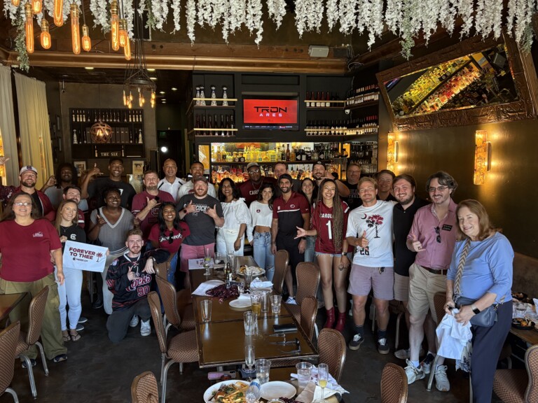 A diverse group of people poses for a photo inside a restaurant, wearing outfits in garnet and black, with some holding a sign that reads 'FOREVER TO THEE'. The restaurant features a stylish interior with plants, bar shelves, and decorative lighting.