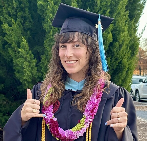 A graduate wearing a black cap and gown adorned with a purple and pink floral lei is smiling and giving a thumbs up gesture with both hands, standing in front of a lush green tree and a parking lot.