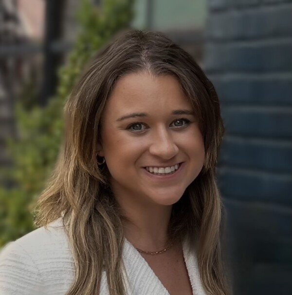 A woman with long, light brown hair and blue eyes smiles warmly at the camera, wearing a white sweater in front of a blurred background featuring green foliage and dark brick.