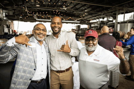 Three men posing for a photo indoors at a gathering, with a casual atmosphere and string lights overhead. One man on the left in a plaid jacket is making a hand sign, while the man in the middle is smiling and gesturing with his hand. The man on the right is wearing a white polo with 'CAROLINA GAMECOCKS' and a red cap, also making a hand sign.
