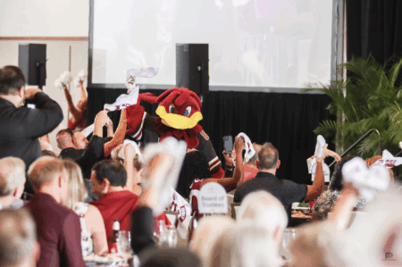 A crowd at a festive event enthusiastically waving white towels, with a red mascot in a sports jersey leading the excitement. In the background, banners and a projector screen are visible, indicating a lively atmosphere.