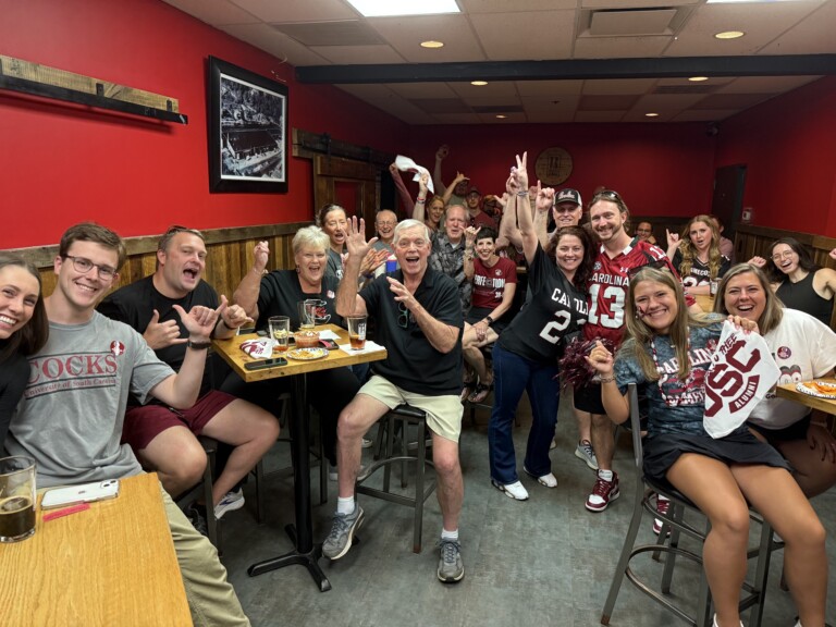 A lively group of fans in a bar, wearing various clothing items representing the University of South Carolina, cheering and celebrating with visible excitement. The setting features a red wall and wooden tables, along with drinks and snacks on the tables.