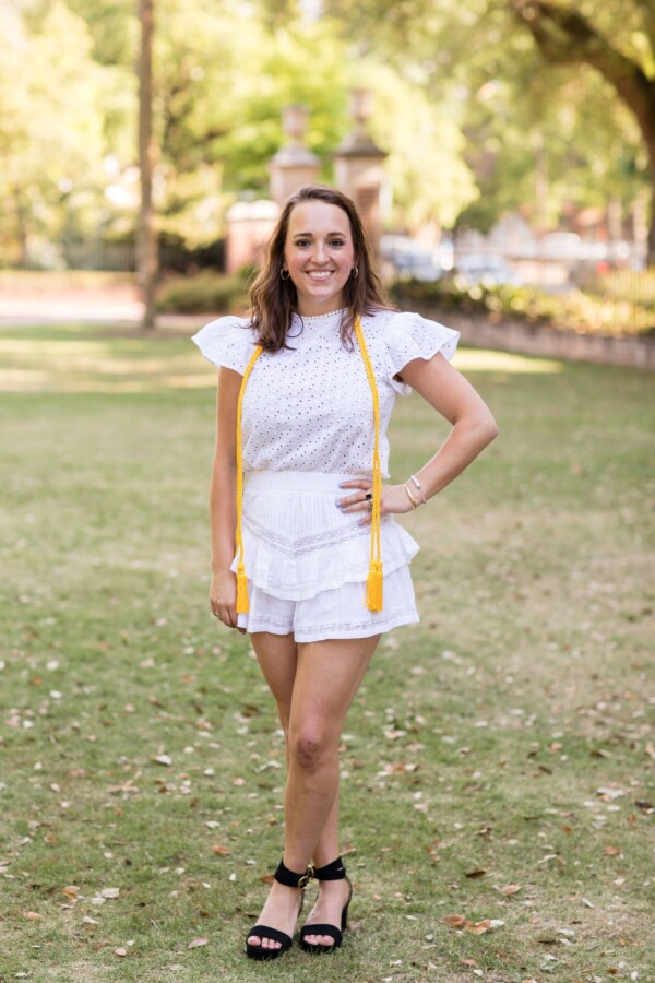A woman stands smiling in a green park, wearing a white eyelet top with puffed sleeves and matching white shorts with ruffles. She accessorizes with a yellow graduation cord draped around her neck and black high-heeled sandals.