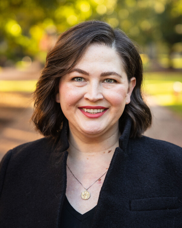 A smiling woman with wavy dark hair, wearing a black coat and a pendant necklace, poses against a blurred natural background with trees and sunlight.