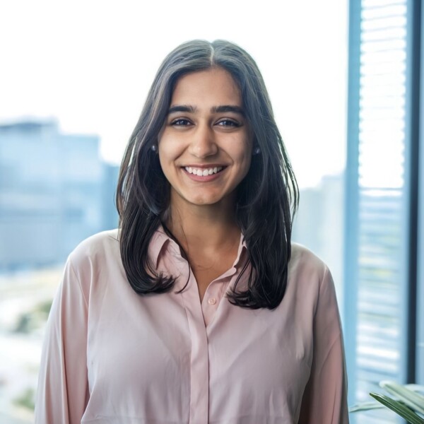 A smiling woman with long black hair wearing a light pink shirt, standing in an office setting with large windows and a blurred cityscape in the background.