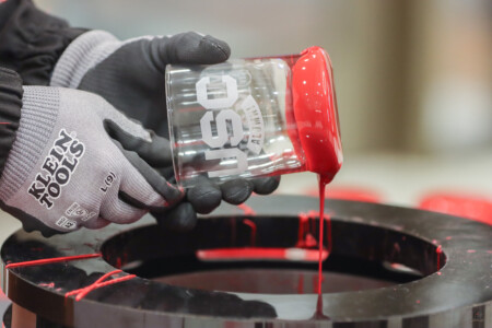 A person wearing gray gloves is tilting a clear glass jar with a red cap, allowing red liquid to pour into a circular black container below. The glass is etched with the text "USC ALUMNI." Red paint drips from the edge of the cap.