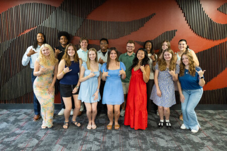 A group of diverse young adults poses together in a brightly lit room with a dark wooden wall design. They display hand signs, with several wearing casual summer outfits in various colors. A woman in a long red dress stands prominently in the center.