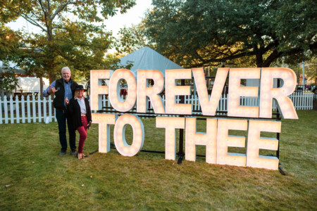 A man and a woman pose in front of large illuminated letters spelling "FOREVER TO THEE" in a grassy area, with trees and a white picket fence in the background.