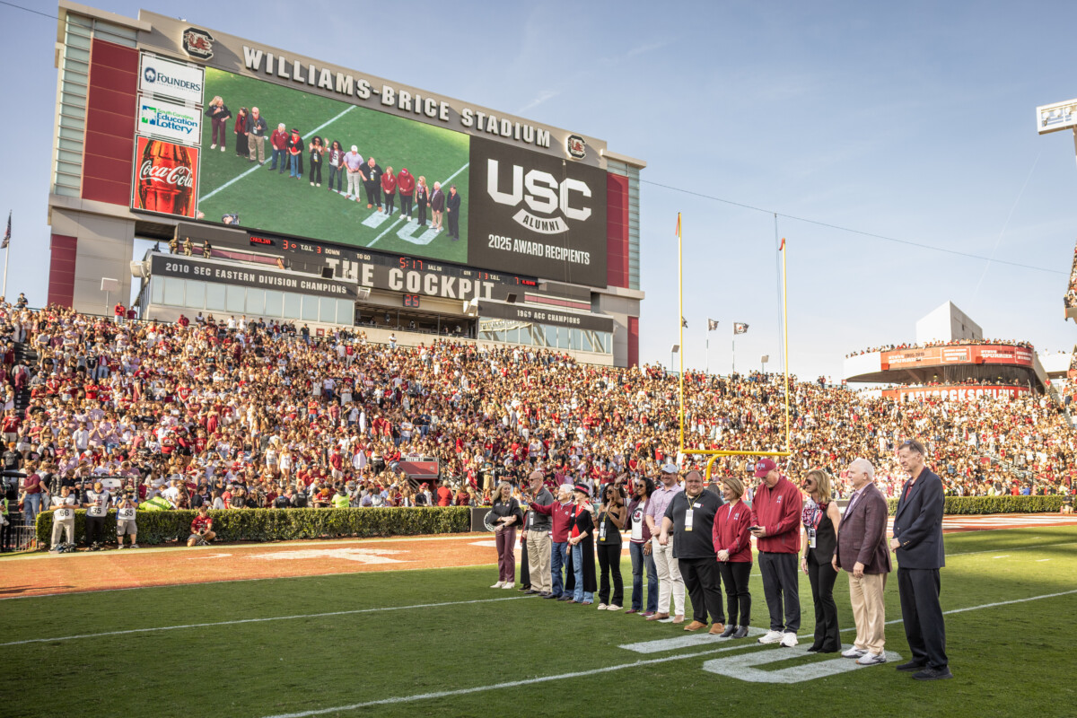 A crowded Williams-Brice Stadium during a football game, featuring a large video screen displaying "USC Alumni 2025 Award Recipients". On the field, a group of individuals stands in recognition, while a cheering crowd fills the seats around them.