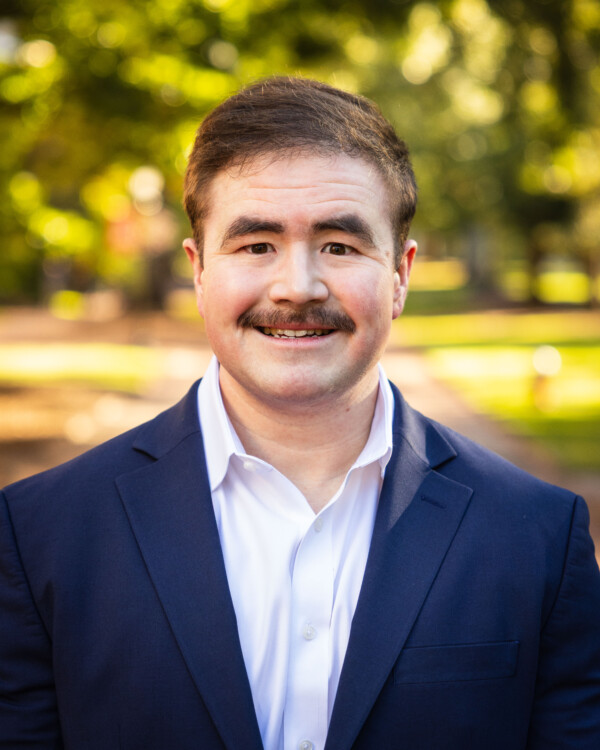 A man in a navy blazer and white dress shirt smiles warmly, standing outdoors with a blurred green background, indicating a park setting.