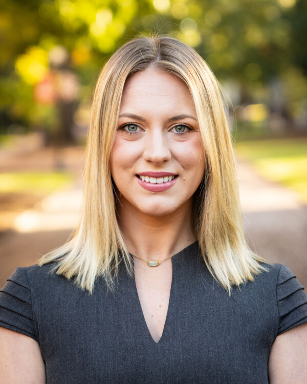 A woman with long, straight blonde hair and green eyes smiles at the camera while wearing a dark gray dress. She is outdoors with a blurred background of greenery and a pathway.