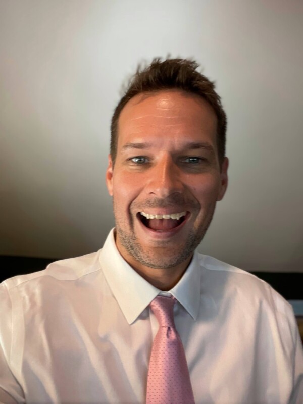 A man smiling enthusiastically, wearing a white dress shirt and a light pink tie with small dots, in a well-lit indoor setting.