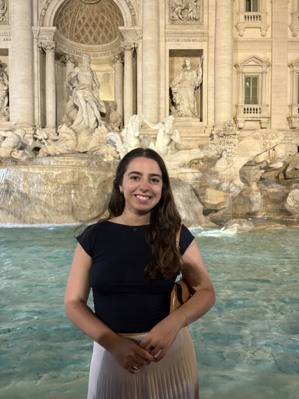 A woman stands smiling in front of the Trevi Fountain at night, with illuminated statues and flowing water in the background. She is wearing a navy blue top and a white pleated skirt, holding a small bag.