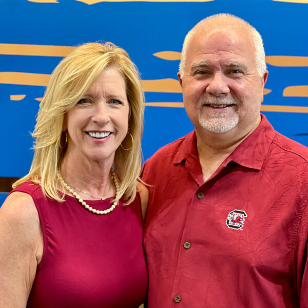 A man and a woman are smiling together. The woman has long blonde hair and is wearing a burgundy dress, along with a pearl necklace. The man has short gray hair and a beard, wearing a burgundy shirt with a design. They are standing in front of a blue background with abstract shapes.