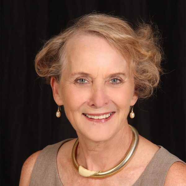 A smiling older woman with short, wavy hair styled loosely, wearing pearl earrings and a chunky gold necklace, posed against a black background.