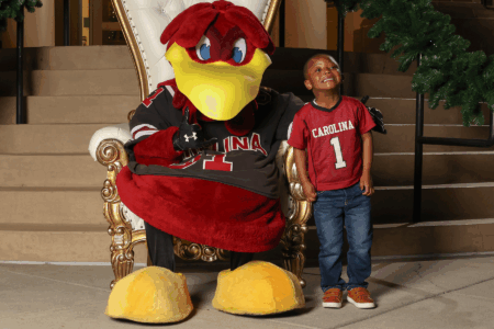 A cheerful young boy stands next to a large, friendly red bird mascot, both seated on an ornate golden chair. The boy wears a red jersey with 'CAROLINA' printed on it, while the mascot wears a dark jersey with 'CAROLINA' and a number. The setting features festive decorations, including garland in the background.