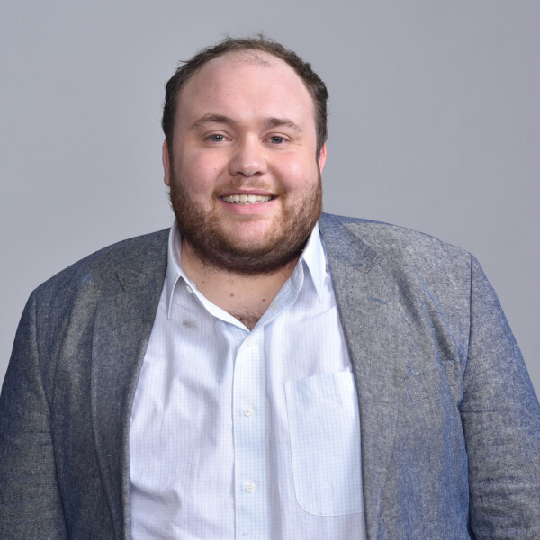 A smiling man with a full beard, wearing a gray blazer over a light checkered shirt, stands against a light gray background.