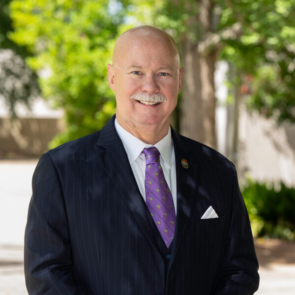 A middle-aged man with a shaved head and white mustache, wearing a black pinstripe suit and a purple tie with small floral designs, stands outdoors in front of a blurred background of green trees and plants, smiling at the camera.