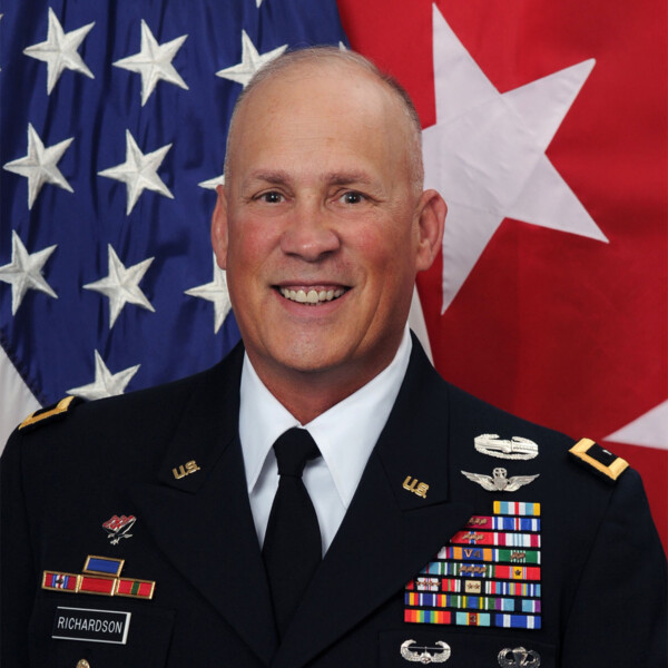 A senior military officer in uniform with various medals and insignia, smiling in front of an American flag and a red flag with a large white star.